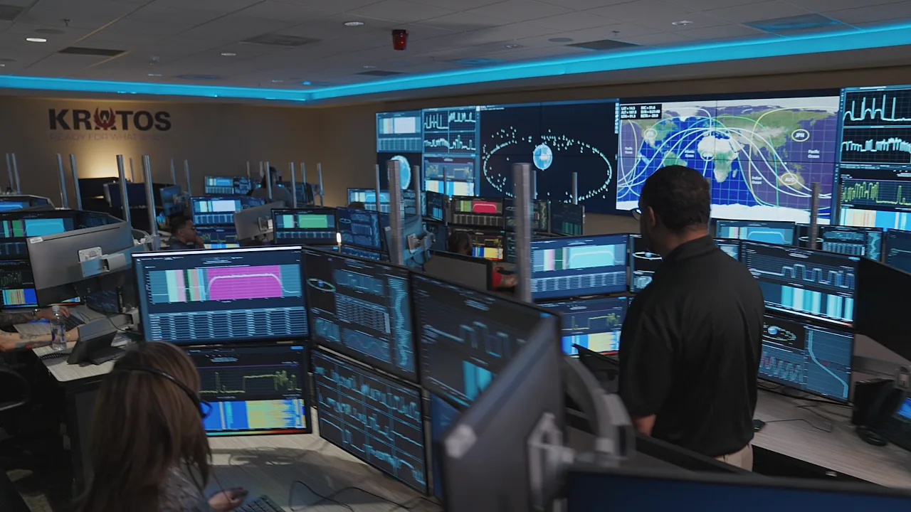 A modern operations center at Kratos Defense with professional technicians monitoring multiple rows of computer monitors displaying advanced data visualization, signal metrics, and global satellite telemetry maps.